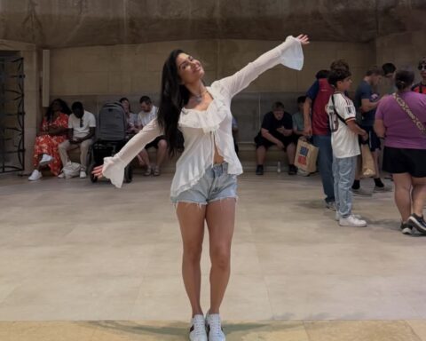Woman in a white blouse and light denim shorts stands with arms stretched, posing as others sit and chat in the background at a public space.
