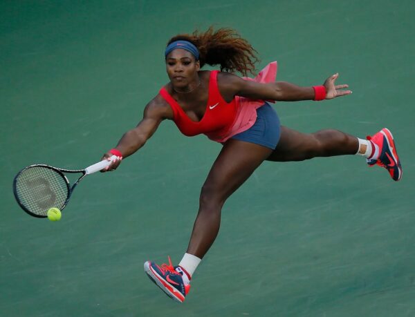 Female tennis player in a red top and blue shorts dives to hit a ball with her racket on a green court, intense focus visible.