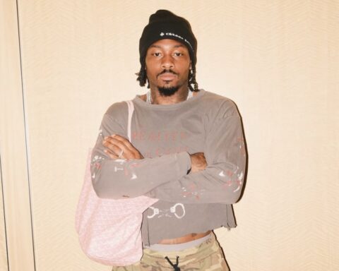 Young man with a black beanie and shoulder bag stands with arms crossed, wearing a paint-splattered gray shirt and camo pants in a neutral background.