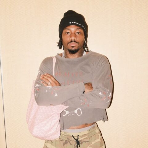 Young man with a black beanie and shoulder bag stands with arms crossed, wearing a paint-splattered gray shirt and camo pants in a neutral background.