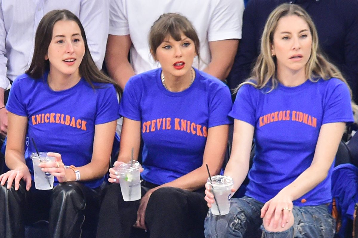 Three women in blue T-shirts with orange text sit on a bench, each holding a clear drink with a straw.