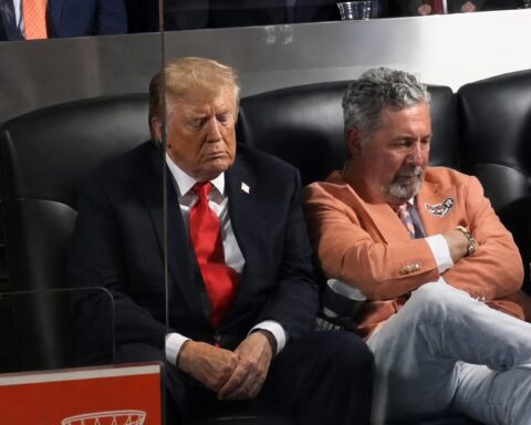Two men sit side by side in black leather seats; the man on the left wears a dark suit with a red tie and has eyes closed in a thoughtful pose.