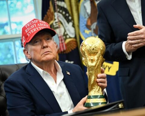 Man in a navy suit sitting at a desk, wearing a red hat that reads 'TRUMP WINS RIGHT ABOUT EVERYTHING,' holding a large gold trophy in one hand with a ceremonial backdrop behind him.