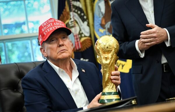 Man in a navy suit sitting at a desk, wearing a red hat that reads 'TRUMP WINS RIGHT ABOUT EVERYTHING,' holding a large gold trophy in one hand with a ceremonial backdrop behind him.