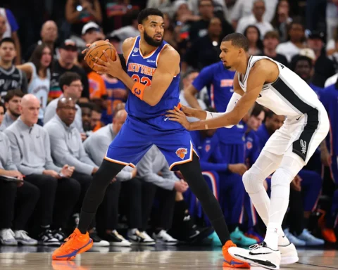 Knicks player in blue drives with the ball as a Spurs defender reaches in on a crowded game court with fans watching.