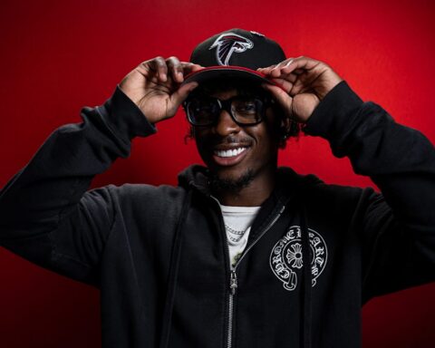 Smiling man wearing a black zip hoodie and glasses, adjusting a cap against a red backdrop.