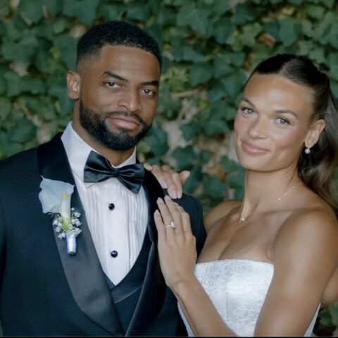 Bride and groom pose in wedding attire outdoors, she shows her engagement ring on her left hand while resting it on his chest.