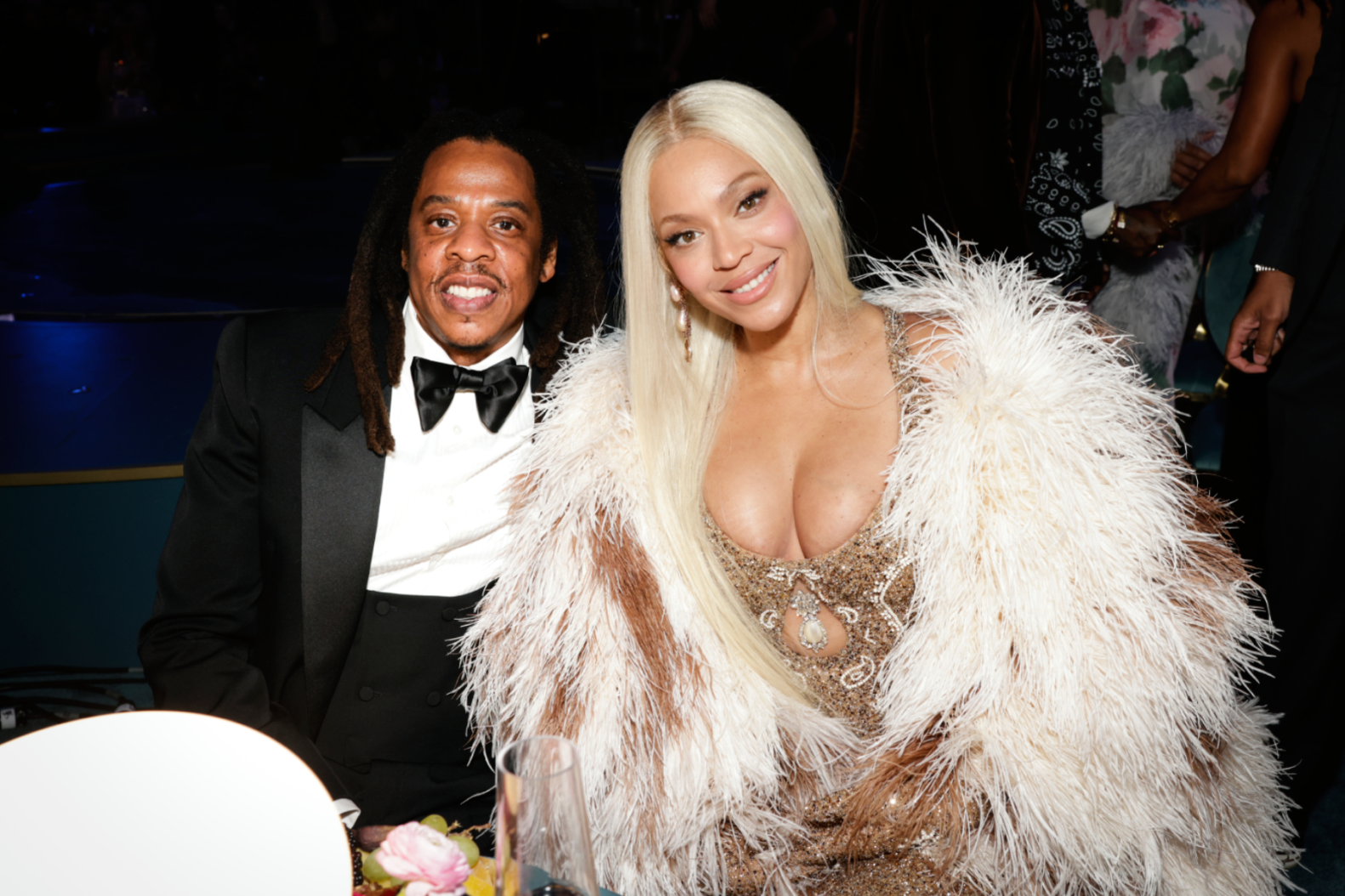 Couple in formalwear posing at a table during a glamorous event; man in black tux, bow tie, woman in sequined dress with large white feathered coat smiles at camera