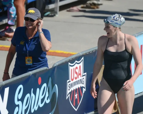 Swimmer in a black one-piece and swim cap smiles as she walks beside a blue barrier banner labeled USA Swimming.