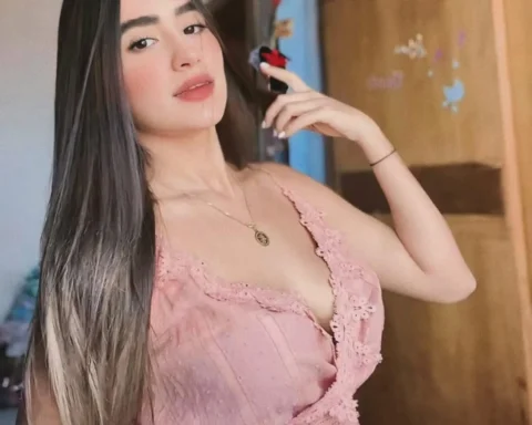 Young woman with long dark hair wearing a pink lace top, posing with her hand near her face indoors.