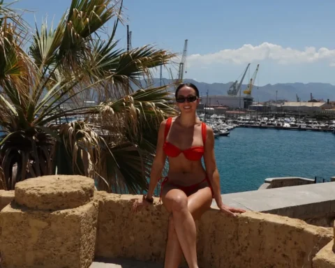 Woman in a red bikini sits on a stone ledge by a harbor, with palm fronds and moored boats in the background under a sunny sky.