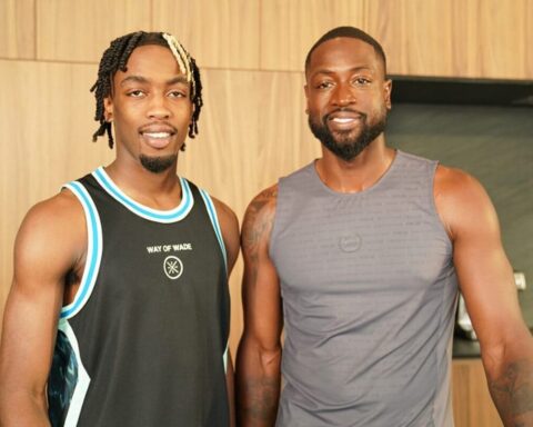 Two smiling men in sleeveless athletic tops posing side by side in a wood-paneled gym area.