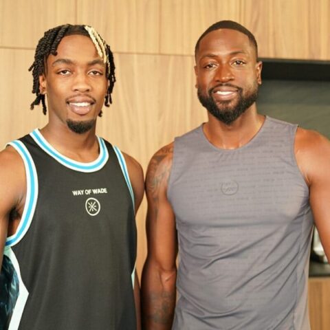 Two smiling men in sleeveless athletic tops posing side by side in a wood-paneled gym area.