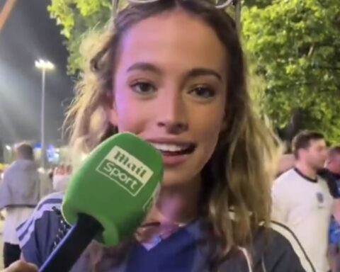 Female reporter speaking into a green mic labeled 'main sport' at an outdoor event, nighttime with trees and people in background