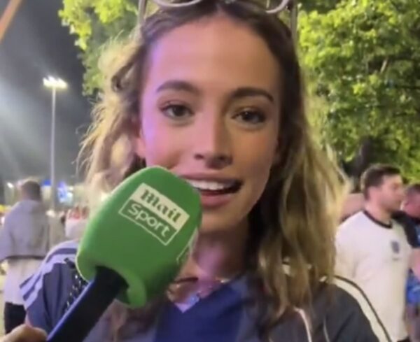 Female reporter speaking into a green mic labeled 'main sport' at an outdoor event, nighttime with trees and people in background