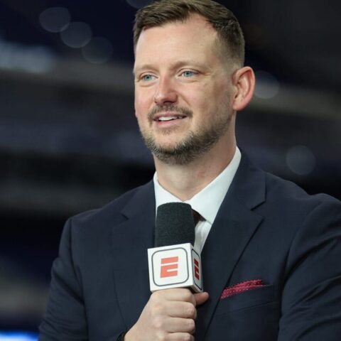Male sports reporter in a dark suit holds a microphone with an ESPN logo during an interview in a studio setting.