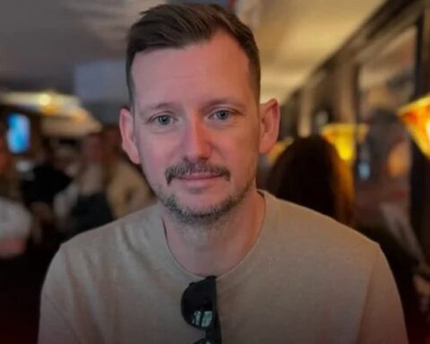 Close-up portrait of a man with short brown hair and stubble, wearing a beige shirt and sunglasses hanging from his neckline, in a dim indoor setting.