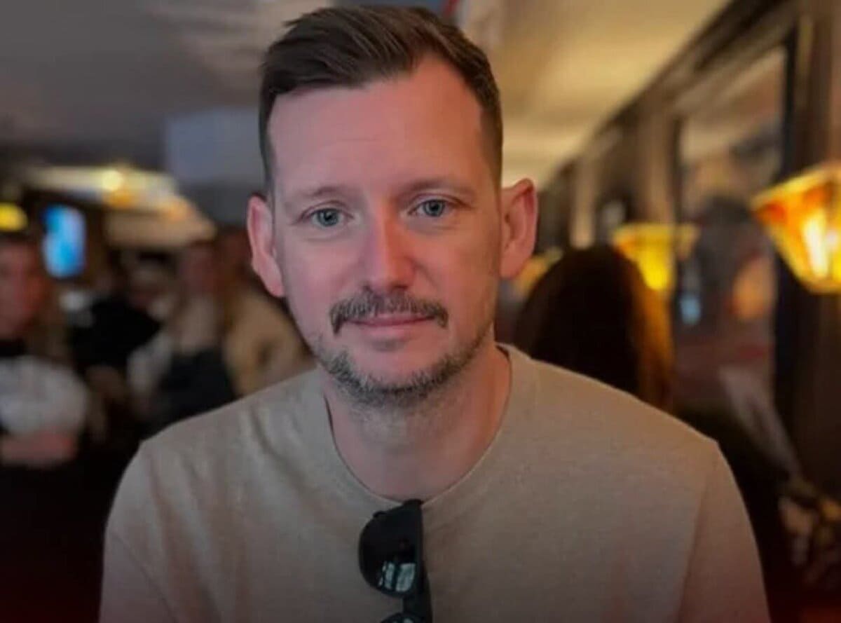 Close-up portrait of a man with short brown hair and stubble, wearing a beige shirt and sunglasses hanging from his neckline, in a dim indoor setting.