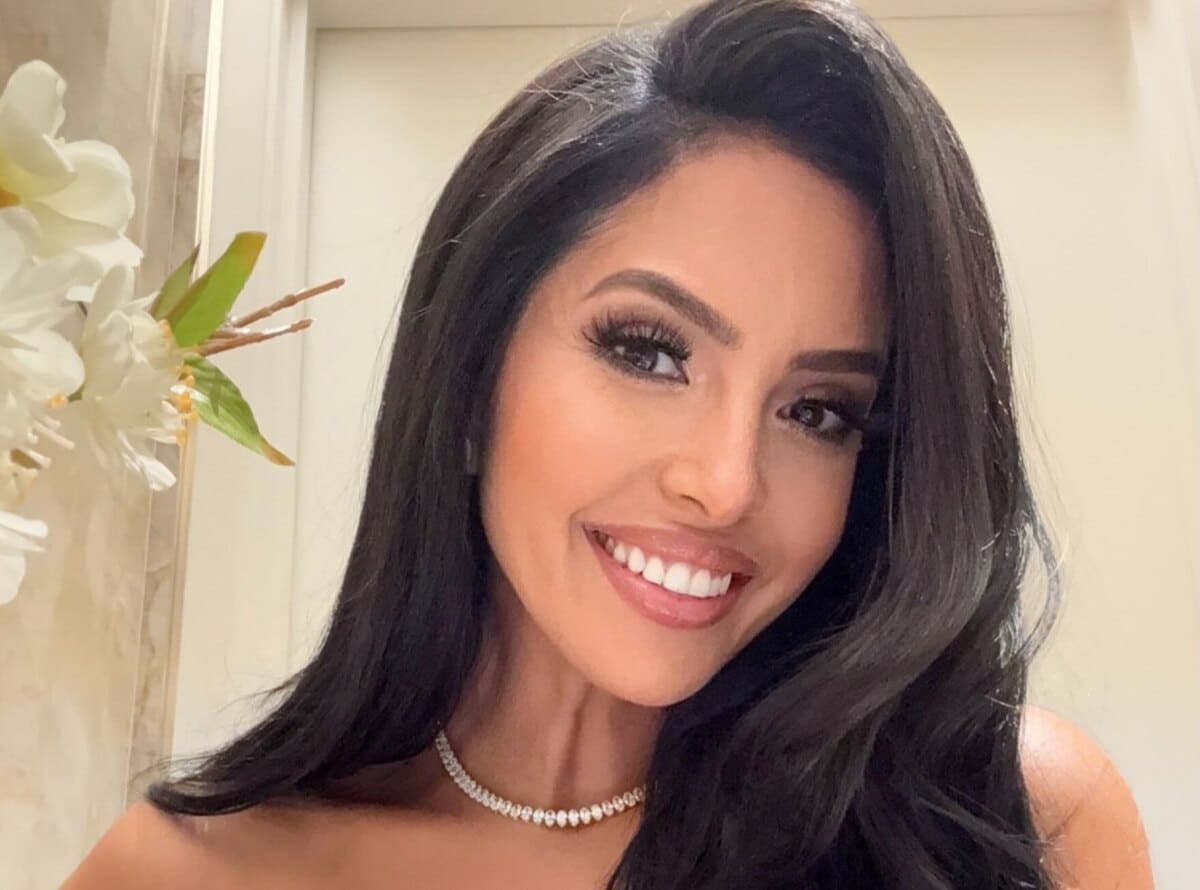 Smiling woman with long black hair wearing a pearl necklace, close-up portrait with white flowers in the background.