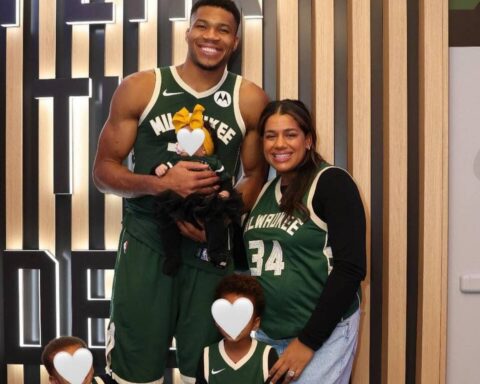 Tall man in a green Milwaukee Bucks jersey stands with a smiling woman, holding a baby with a yellow bow; two children in front wear matching jerseys, posing indoors.