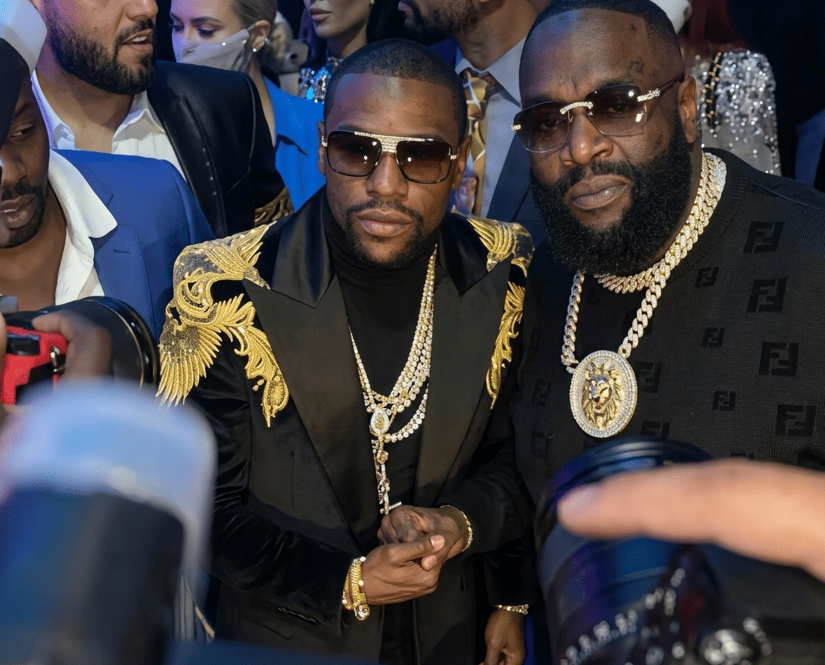 Two men in flashy attire pose for photos at a crowded event: one with a black and gold embroidered jacket and layered gold chains, the other in a black Fendi-knit top with a large gold medallion; several photographers and people surround them.