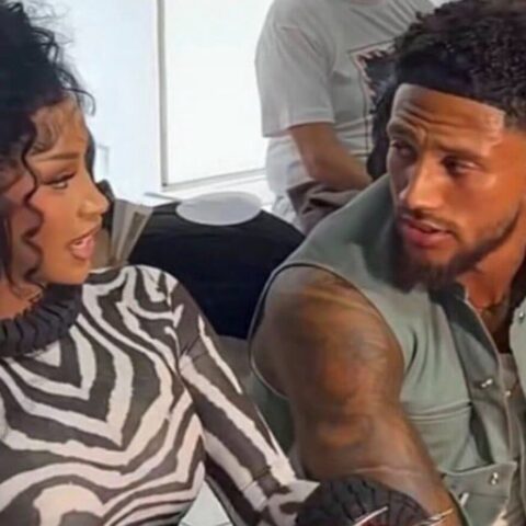 Woman with curly hair in a zebra-print turtleneck talking to a bearded man with tattoos on his arm, seated indoors and looking at each other.