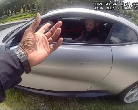 Person with a dark sleeve raises a hand toward a silver car window as the driver looks surprised; grassy roadside with guardrail in background.