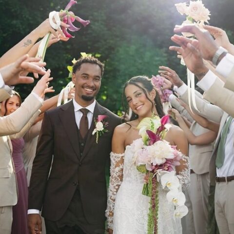Bride and groom walking through a celebratory aisle, guests in beige outfits cheer with flowers and ribbons overhead.