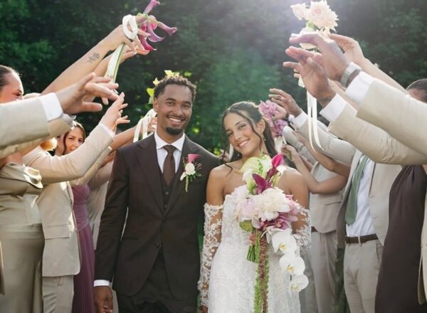 Bride and groom walking through a celebratory aisle, guests in beige outfits cheer with flowers and ribbons overhead.