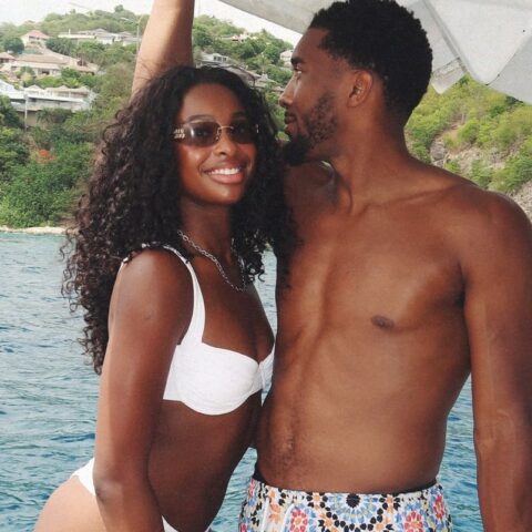 Smiling couple in swimwear on a boat, with blue water and rocky, green hills in the background.