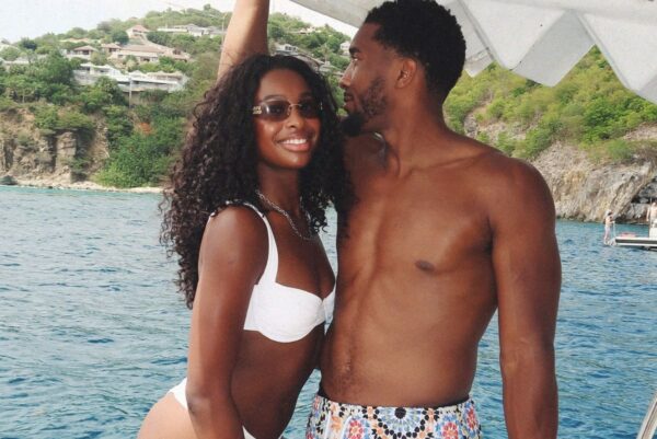 Smiling couple in swimwear on a boat, with blue water and rocky, green hills in the background.
