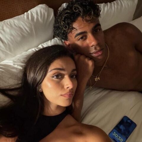 Couple lying on a bed together; woman in front taking a selfie while a shirtless man rests beside her wearing gold chains.