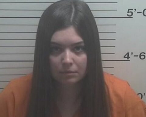 Mugshot of a young woman in an orange jumpsuit standing against a height chart, looking at the camera.