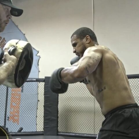 Boxer practicing punches with a trainer holding focus mitts inside a training ring in a gym cage.
