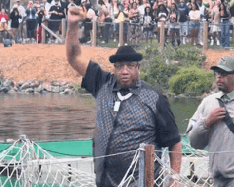 Man in a black hat and checkered vest raises his right fist at a waterfront event, crowd behind him.