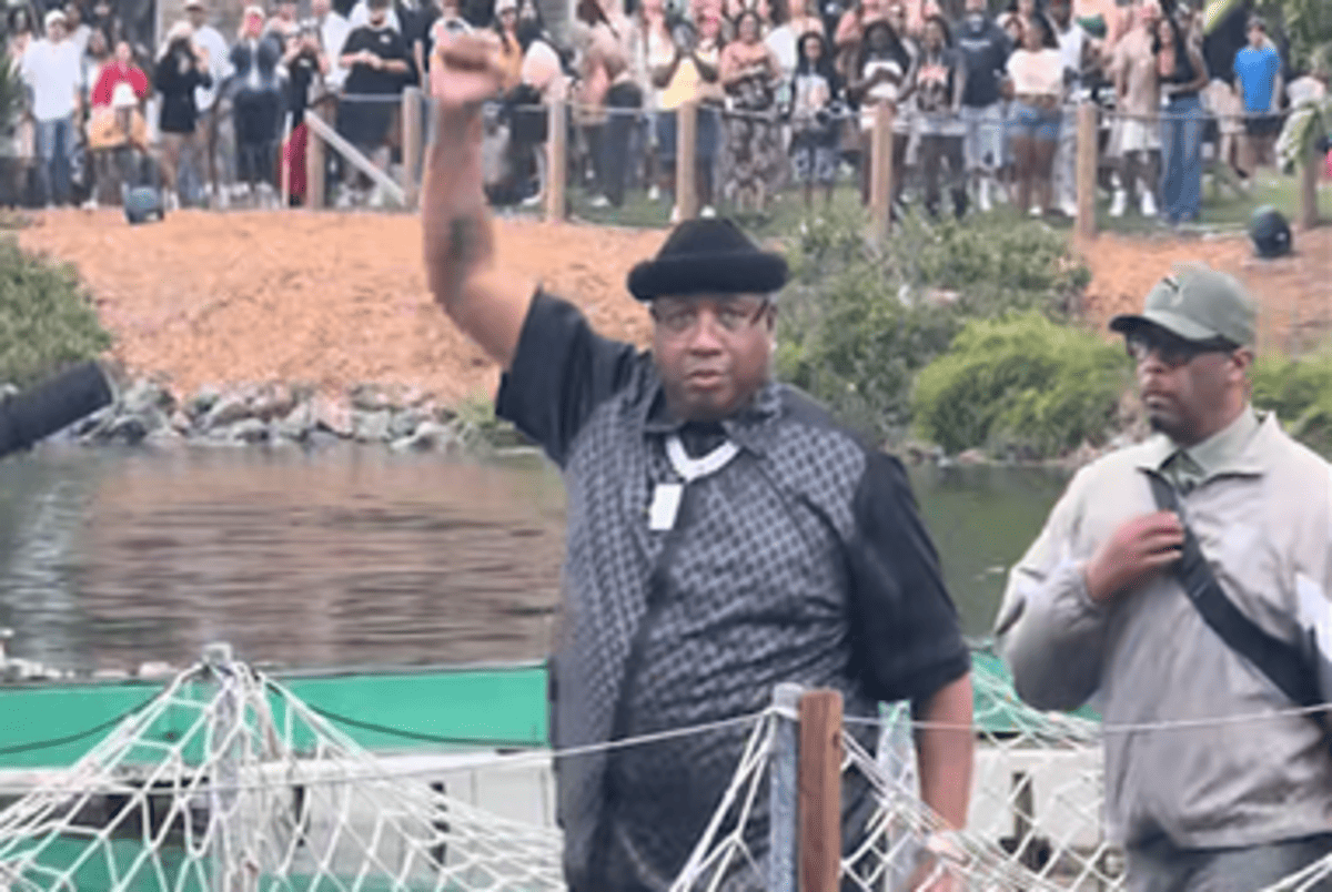 Man in a black hat and checkered vest raises his right fist at a waterfront event, crowd behind him.