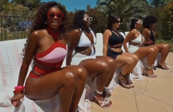 Six women in stylish swimsuits posing on a poolside bench in bright sunlight, wearing sunglasses and heels/ sandals.