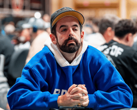 Man with a beard in a blue hoodie and brown cap sits at a crowded table, hands clasped, looking thoughtful at an indoor event.