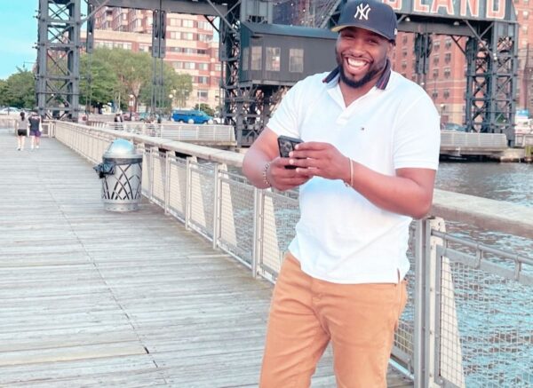 Smiling man in a white polo and tan shorts stands on a waterfront boardwalk, looking at his phone by the railing.