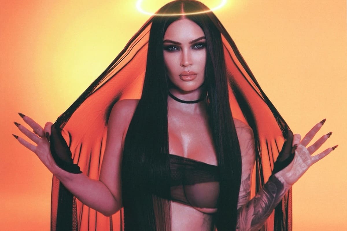 Portrait of a woman with long black hair, wearing a sheer black top and choker, holding orange fabric around her shoulders with a glowing halo effect above her head against an orange backdrop.