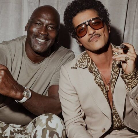 Two men posing for a fashion photo, one in a beige tee with camo pants and the other in a tan suit with leopard-print shirt and sunglasses, both holding cigars and smiling.