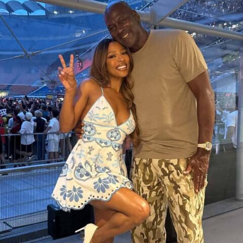 Happy couple posing for a photo at a crowded indoor event; woman in a blue-and-white floral dress makes a peace sign beside a tall man in a tan shirt and camo shorts.