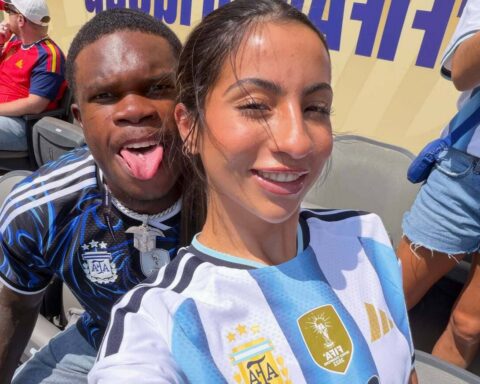 Two friends pose for a selfie in stadium seats; woman in an Argentina football jersey smiles, man behind her sticks out his tongue.