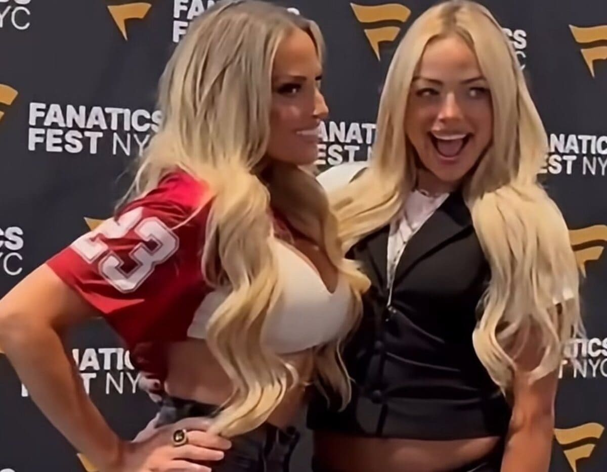 Two smiling women pose for a photo at a Fanatics Fest NY backdrop; one wears a red football jersey and white top, the other a black blazer.