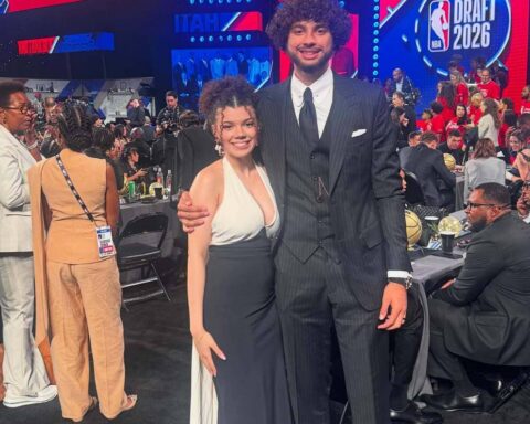 Tall man in a dark pinstripe suit and tie stands with a smiling woman in a white halter top and black skirt at a crowded formal event, with bright NBA Draft 2026 screens in the background.