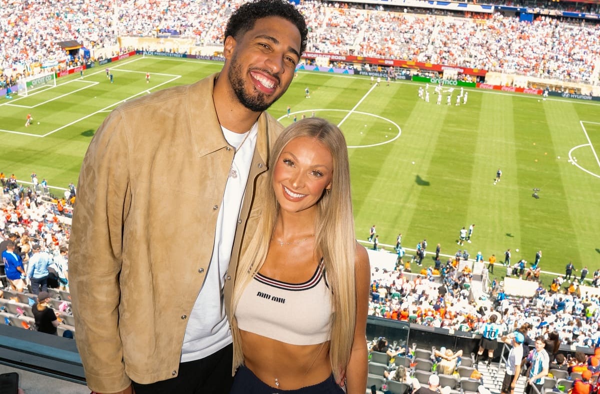 Smiling couple posing for a photo in a soccer stadium with a green pitch and cheering spectators in the stands.