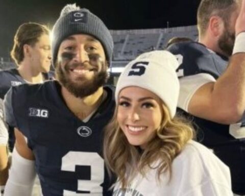 Smiling man in a dark football jersey with face paint and a beanie stands beside a woman in a white beanie at a stadium.