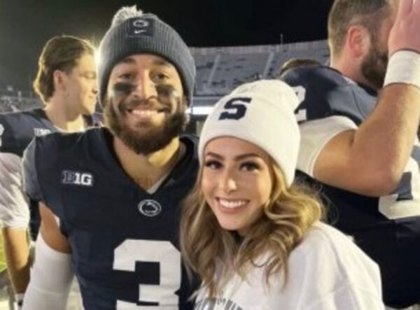 Smiling man in a dark football jersey with face paint and a beanie stands beside a woman in a white beanie at a stadium.