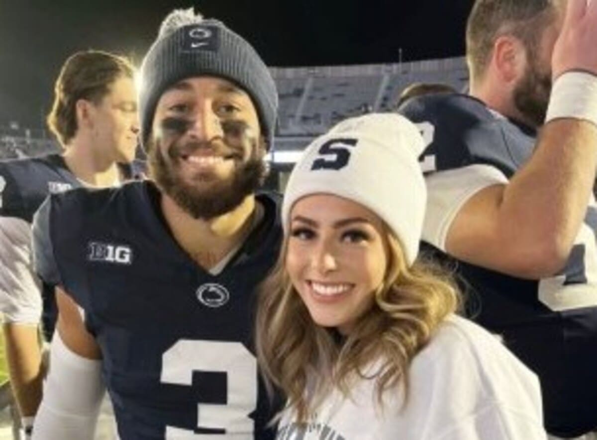Smiling man in a dark football jersey with face paint and a beanie stands beside a woman in a white beanie at a stadium.