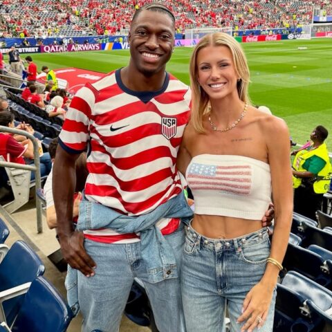 Smiling couple poses together in a stadium: man in a red-and-white USA jersey with denim jacket tied around the waist, woman in a white crop top with a US flag, both wearing jeans; green field behind them.